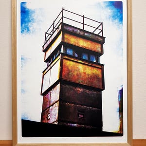 Op de afbeelding: Een vintage foto van een hoge, meerlaags betonnen wachttoren met een metalen railing op de top. De toren is geschilderd in tinten geel, oranje en bruin, met een vervaagde blauwe lucht op de achtergrond.