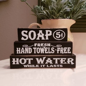 May include: Three black wooden signs stacked on top of each other. The top sign says "SOAP 5¢" in white letters. The middle sign says "FRESH HAND TOWELS-FREE" in white letters. The bottom sign says "HOT WATER WHILE IT LASTS" in white letters.