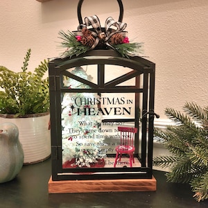 May include: Black metal lantern with a wooden base, decorated for Christmas. The lantern features a small Christmas tree and a red chair inside. The top is adorned with a plaid bow, pine needles, and pine cones. Text reads "Christmas in Heaven."