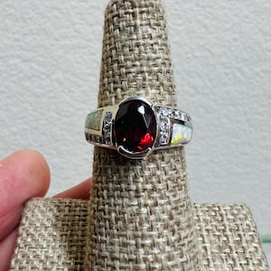 May include: A silver ring featuring a large, oval-cut, deep red gemstone as the centerpiece. The ring is flanked by smaller, white gemstones and opal-like stones on either side. The ring is displayed on a textured, beige ring holder.