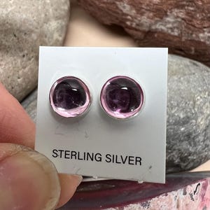 May include: A pair of sterling silver stud earrings with pink glass cabochons. The earrings are displayed on a white card with the words "Sterling Silver" printed on it.