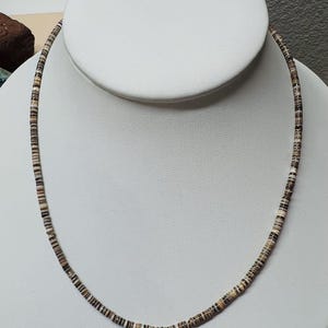 May include: A delicate necklace composed of small, flat, circular beads. The beads are primarily neutral tones of brown, tan, and black, strung together to form a single strand. The necklace is displayed on a white bust.