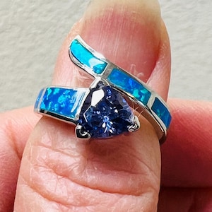 May include: A silver ring with a triangular, dark blue gemstone at the center. The ring band features blue opal inlays. A second band of blue opal crosses over the main ring. The ring is shown on a finger.