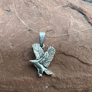 May include: A silver-toned metal pendant shaped like an eagle in flight, featuring detailed wing feathers and a sharp beak. The pendant is suspended from a thin, clear, flexible monofilament cord that loops through a small silver-toned metal bail.