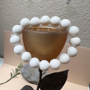 May include: A white beaded bracelet with round beads, displayed on a decorative stand with a brown base and black metal leaves.