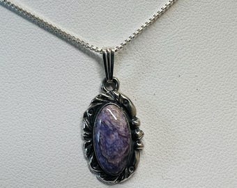 May include: A silver pendant necklace featuring an oval, purple gemstone. The gemstone is set in a decorative silver frame with a detailed design. The necklace has a delicate silver chain.