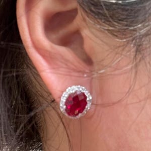 May include: A pair of red gemstone earrings with a silver halo setting. The earrings are worn in a person's ear.
