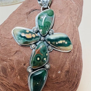 May include: A silver pendant in the shape of a cross, made with five pieces of green, brown, and white patterned stone. The pendant is set on a brown rock.