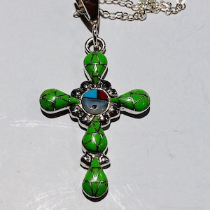 May include: A silver necklace with a green cross pendant. The cross is decorated with turquoise and other stones. The pendant is attached to a silver chain.