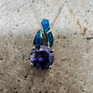 May include: A silver necklace with a pendant featuring a large, round, purple gemstone. The pendant has a decorative top with blue and green accents. The necklace is displayed on a textured, gray surface.