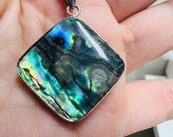 May include: Abalone shell pendant necklace. The pendant is a square shape with a silver-colored bezel. The abalone shell has iridescent colors of blue, green, and purple. The necklace chain is silver-colored.