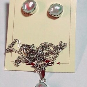 May include: A set of pearl jewelry featuring a necklace and earrings. The necklace has a silver chain and a pendant with a large, iridescent pearl. The earrings are silver studs with smaller, iridescent pearls.