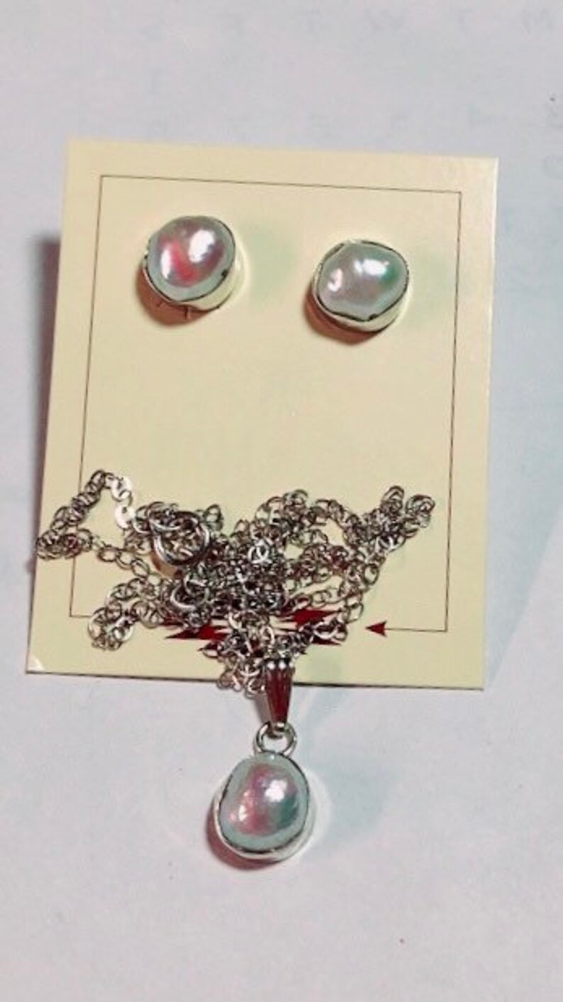 May include: A set of pearl jewelry featuring a necklace and earrings. The necklace has a silver chain and a pendant with a large, iridescent pearl. The earrings are silver studs with smaller, iridescent pearls.