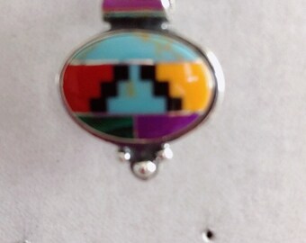 May include: A silver pendant necklace with a turquoise, red, yellow, purple, black, and green stone inlay. The pendant is oval shaped and has a geometric design.