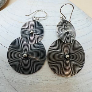 May include: A pair of silver earrings with two circular discs, each with a concentric circle design. The discs are connected by a small silver bead.