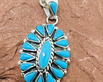 May include: A silver pendant necklace with a turquoise stone design. The pendant is oval shaped and features a cluster of turquoise stones in a sunburst pattern.