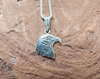 May include: A silver pendant necklace with a detailed eagle head. The pendant hangs from a fine silver chain. The eagle head is intricately carved, showing feathers and a sharp beak. The necklace is displayed on a textured, brown surface.