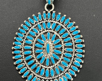 May include: A silver pendant necklace with a turquoise inlay design. The pendant is round and features a circular pattern of turquoise stones.