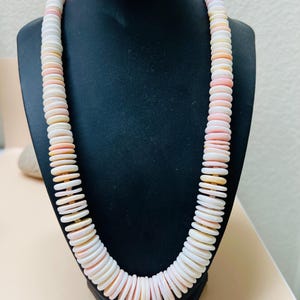 May include: A white, pink, and gold necklace made of round, flat beads. The necklace is displayed on a black mannequin.