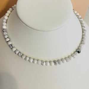 May include: A white howlite beaded necklace on a white display stand. The beads are round and have a marbled pattern.