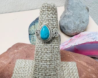 May include: A silver ring with a teardrop-shaped turquoise stone. The ring has a detailed, textured band with spiral accents on either side of the stone. The ring is displayed on a woven ring holder, with rocks in the background.