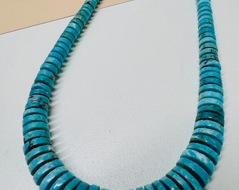 May include: A long necklace composed of numerous small, flat, circular turquoise beads. Each bead features a vibrant cyan blue colour with natural dark brown matrix veining, strung together to form a continuous strand of uniform, disc-shaped stones.