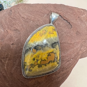 May include: A large, polished pendant with a silver bail. The pendant features a striking, natural stone with a mix of yellow, black, and brown colors. The stone is set in a silver frame with a rope-like border, creating a unique jewelry piece.