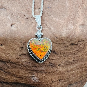 May include: A silver heart-shaped pendant necklace with a colorful, speckled stone center. The stone features shades of orange, yellow, and green. The pendant is framed with a silver rope design and hangs from a delicate silver chain.