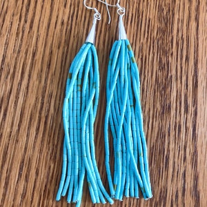 May include: A pair of turquoise beaded earrings with silver-colored metal accents. The earrings feature a fringe design with multiple strands of small, rectangular beads. The beads are a bright turquoise color with occasional brown accents.