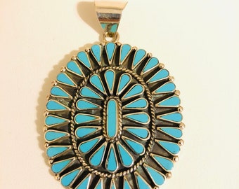 May include: Oval turquoise pendant with a silver setting. The pendant features a central turquoise rectangle surrounded by a double row of turquoise teardrop shapes. The pendant has a silver bail for hanging.