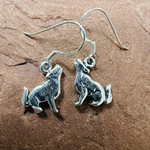 May include: A pair of silver-colored earrings featuring a howling wolf design. The earrings have a hook closure and are displayed on a brown, textured surface. The wolf pendants are detailed with visible fur and facial features.