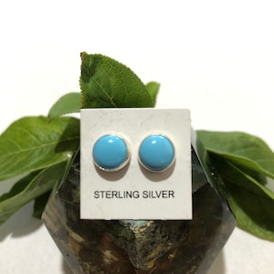 May include: A pair of round, light blue stud earrings with a silver border. The earrings are displayed on a white card that reads "STERLING SILVER". The background includes green leaves and a dark, textured surface.
