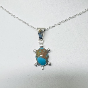 May include: A silver necklace featuring a turtle-shaped pendant. The turtle shell is inlaid with a piece of turquoise stone displaying brown matrix patterns. The pendant hangs from a delicate silver chain, and the bail includes a small turquoise accent.