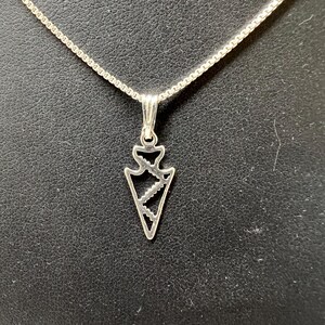 May include: A silver-colored necklace featuring a pendant shaped like an arrowhead. The pendant has a cut-out design with a zig-zag pattern inside. The chain is a delicate, square-link style. The background is a dark, textured surface.