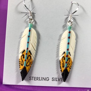 May include: A pair of sterling silver earrings featuring hand-painted bone feather designs. The feathers are white with black, yellow, blue, and red accents. The earrings are displayed on a white card with the text "STERLING SILVER".