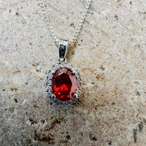 May include: A silver chain necklace with a pendant featuring a large red oval gemstone surrounded by smaller clear gemstones.