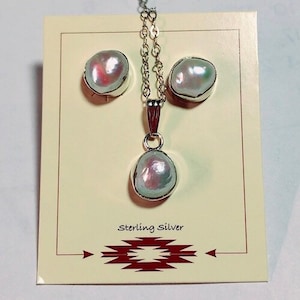 May include: A white pearl necklace and earrings set with a sterling silver chain and pendant. The pearls are iridescent and have a slightly pink hue. The set is displayed on a card with the words "Sterling Silver" printed on it.