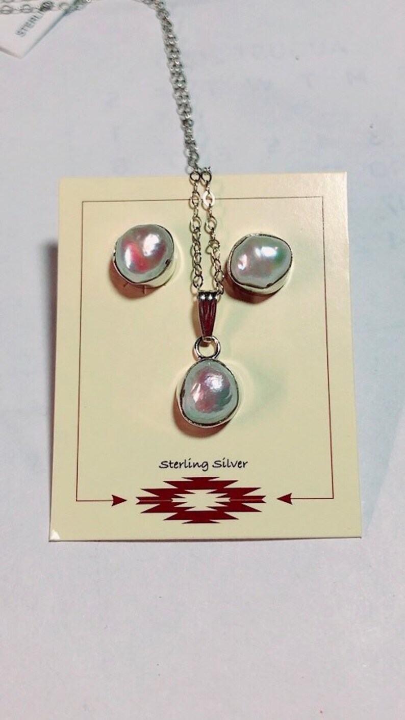 May include: A white pearl necklace and earrings set with a sterling silver chain and pendant. The pearls are iridescent and have a slightly pink hue. The set is displayed on a card with the words "Sterling Silver" printed on it.