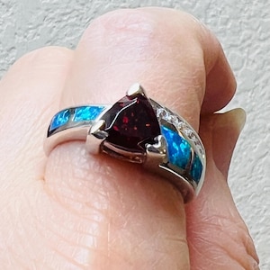 May include: A silver ring featuring a triangular, dark red gemstone at its center. The ring is accented with blue and white stones, creating a striking contrast. The ring is held up by a person's hand.