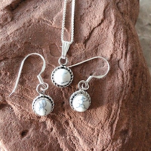 May include: A silver necklace and earrings set with white howlite stone pendants. The pendants are round and have a silver bezel setting. The necklace has a delicate chain.