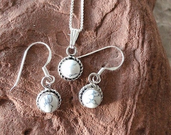 May include: A silver necklace and earrings set with white howlite stone pendants. The pendants are round and have a silver bezel setting. The necklace has a delicate chain.
