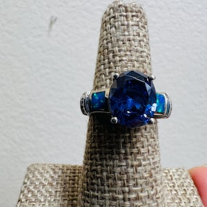 May include: A silver ring featuring a large, oval-cut, deep blue gemstone as the centerpiece. Two smaller, rectangular blue-green stones flank the main stone. The ring is displayed on a textured, tan ring holder.
