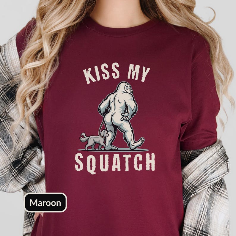 May include: Maroon t-shirt with the text "KISS MY SQUATCH" above a cartoon illustration of a Bigfoot walking a dog. The Bigfoot is white, and the dog is gray. The shirt is a solid maroon color.