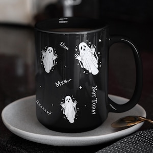 May include: A black ceramic mug featuring three white ghost illustrations and the text "Ugh," "Meh...", "Not Today," and "Really?" printed in white. The mug has a curved handle and is decorated with small white star accents scattered around the ghosts.