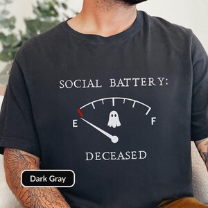 May include: A dark gray t-shirt featuring the white text SOCIAL BATTERY: DECEASED. Below the text is a white fuel gauge graphic with a red indicator at the empty mark, a white ghost icon in the center, and the letters E and F on either side.