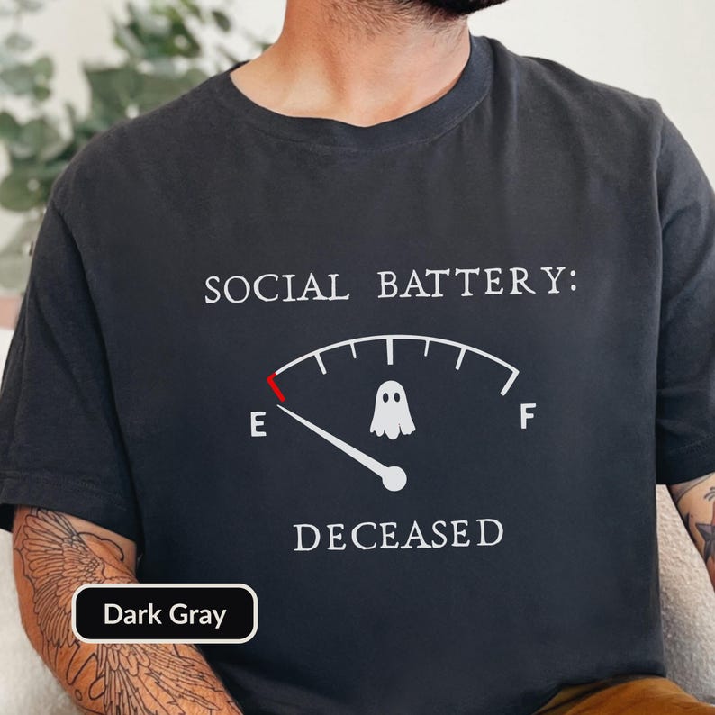 May include: A dark gray t-shirt featuring the white text SOCIAL BATTERY: DECEASED. Below the text is a white fuel gauge graphic with a red indicator at the empty mark, a white ghost icon in the center, and the letters E and F on either side.