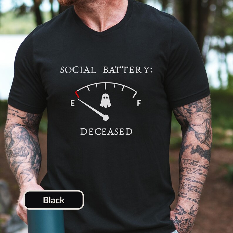 May include: A black V-neck t-shirt featuring a white graphic of a fuel gauge with the text SOCIAL BATTERY: DECEASED. The gauge needle points to E, and a small white ghost icon is positioned in the center of the arc between the E and F markers.