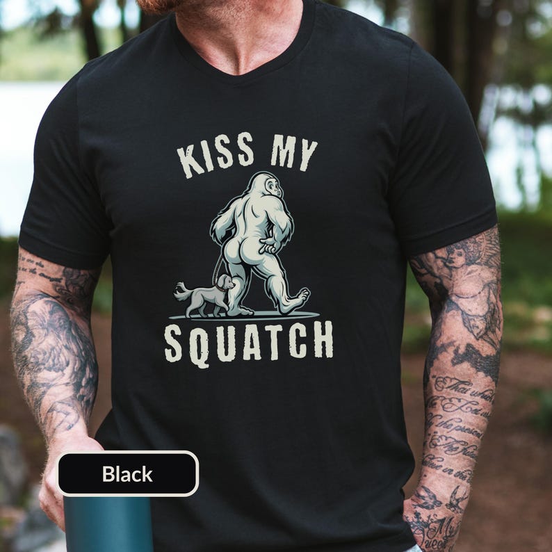 May include: Black t-shirt with the text "KISS MY SQUATCH" above an illustration of a Bigfoot figure walking with a small dog. The Bigfoot is light gray, and the dog is gray. The text is in a white, bold font.