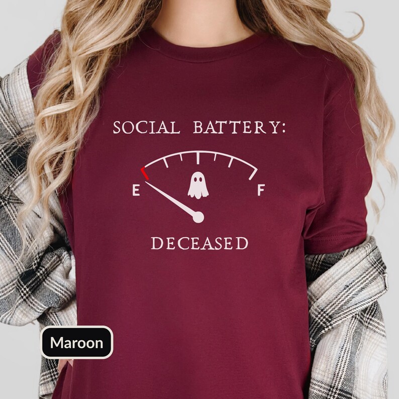 May include: A maroon t-shirt featuring a white graphic of a fuel gauge labeled SOCIAL BATTERY: DECEASED. The gauge needle points to the E, and a small white ghost icon is positioned in the center of the arc, replacing the typical fuel indicator.