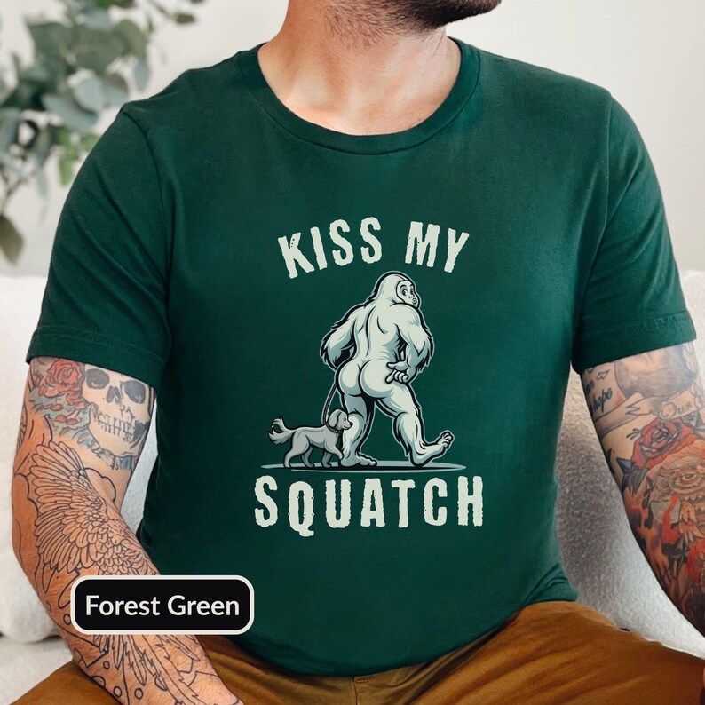 May include: Forest green t-shirt with a graphic of Bigfoot walking a dog, and the text "KISS MY SQUATCH" in white. The shirt is a crew neck style.